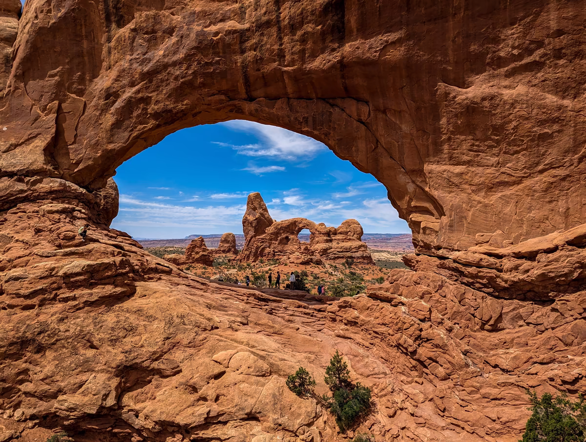 Looking through the giant opening of a red sandstone archway at Arches National Park, revealing a rugged canyon landscape, smaller rock towers, and a group of distant visitors.
