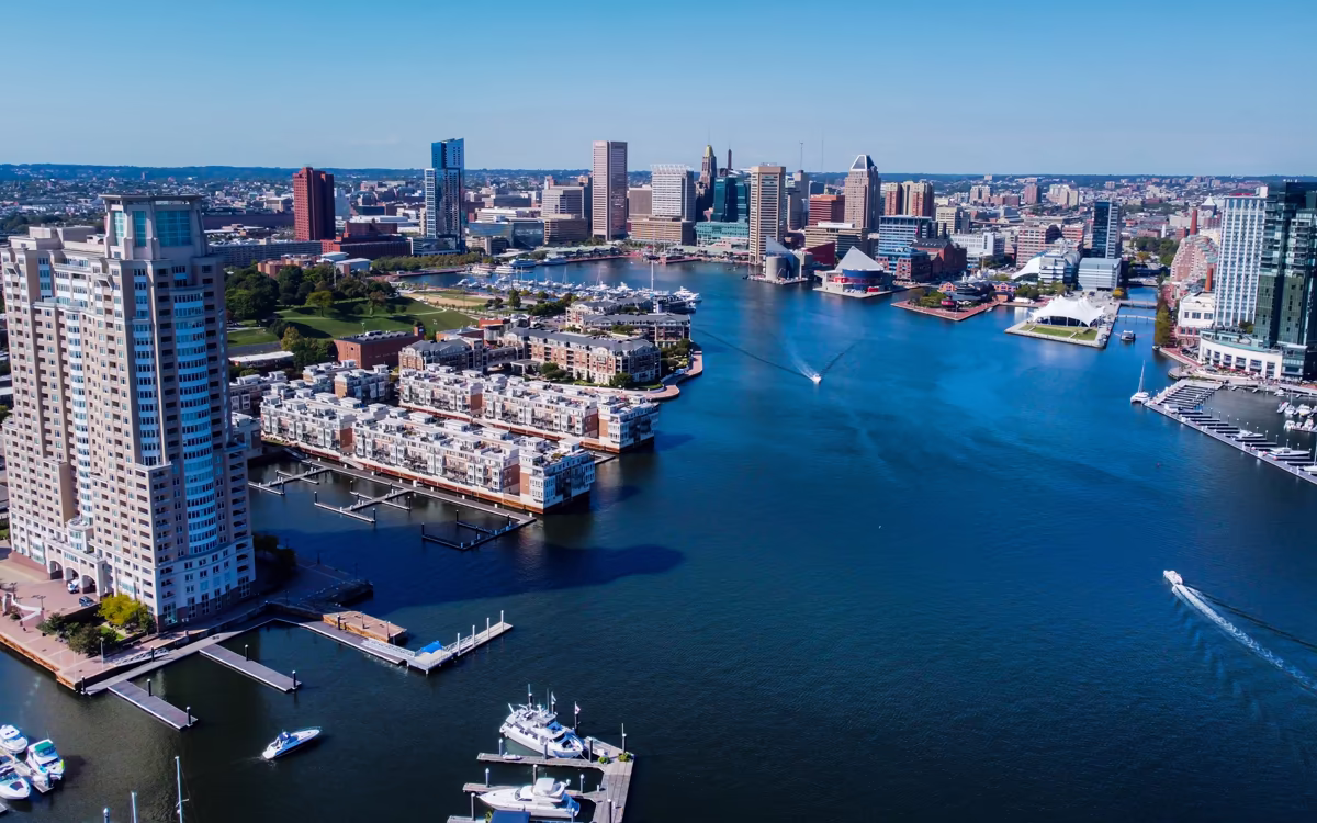 A high-angle drone shot capturing the blue waters of Baltimore's Inner Harbor, featuring speedboats, boat slips, and the distinct city skyline under a clear sky.