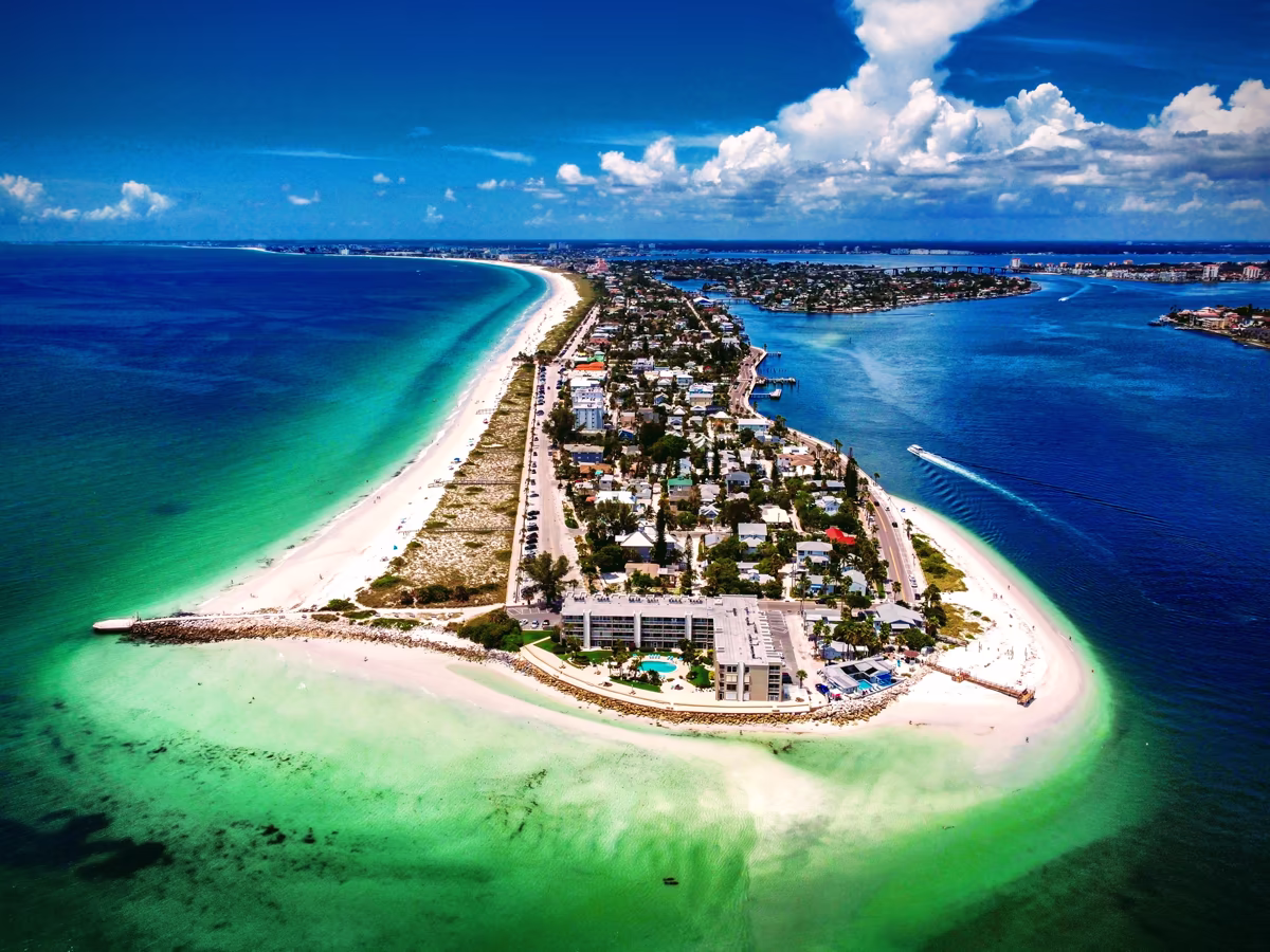 A sweeping overhead view of a coastal peninsula, showing tropical turquoise ocean waters on one side, a white sandy beachfront, coastal properties, and a deep blue bay on the other.