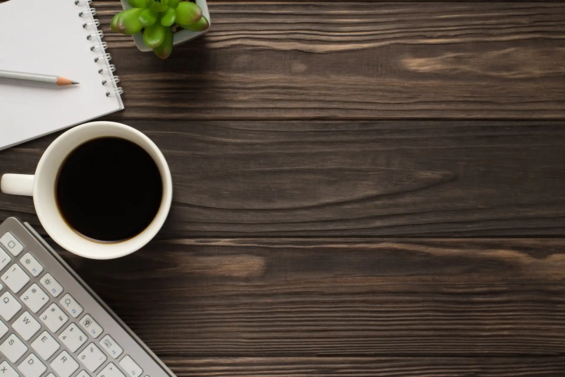 Top-down view of a dark wooden desk with a cup of black coffee, a computer keyboard, a spiral notebook with a pencil, and a small succulent plant, leaving ample copy space on the right.