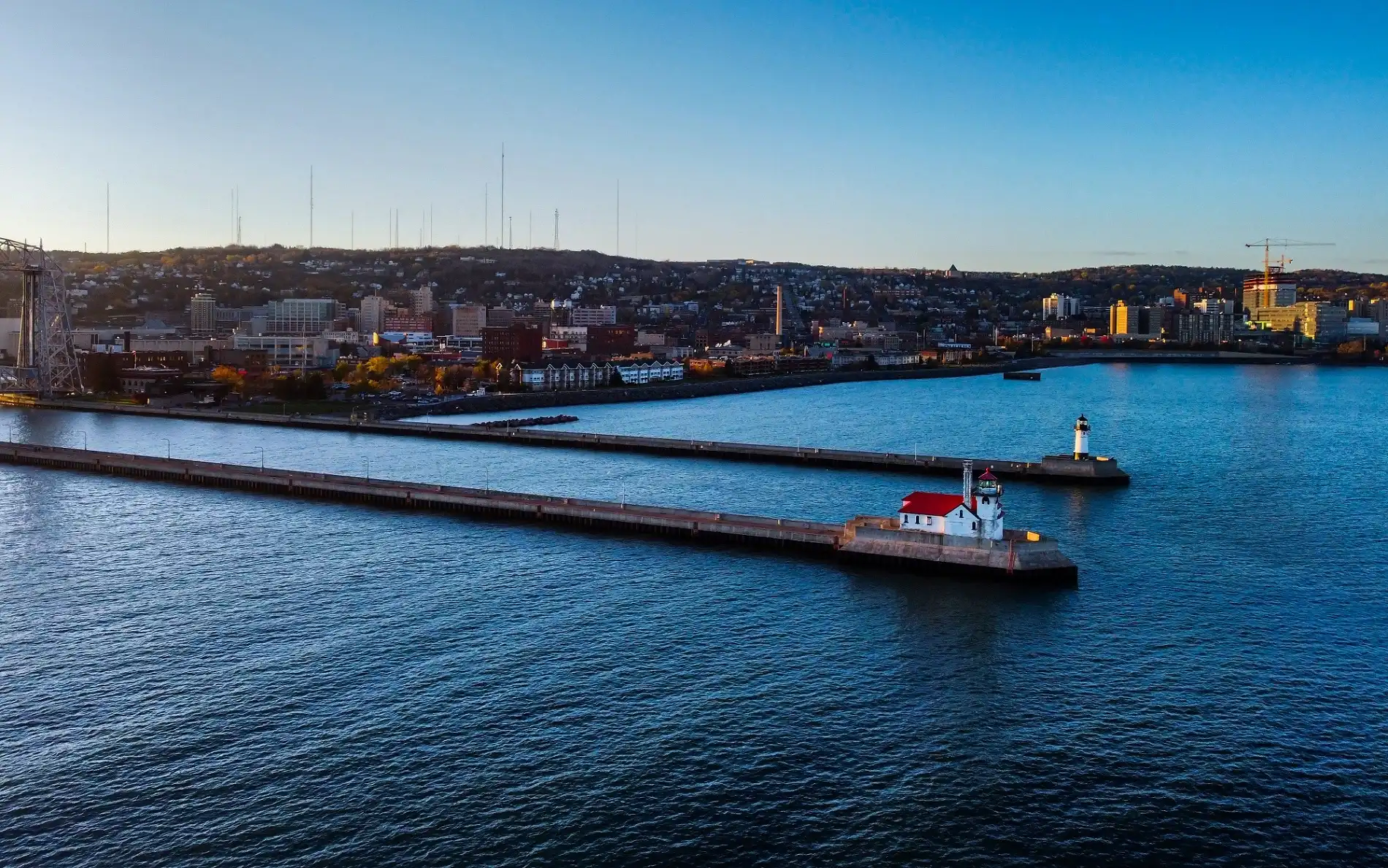An elevated drone perspective of harbor entry piers featuring a white lighthouse with a red roof on the main breakwater, looking across blue water toward a coastal city built onto a rising hill.