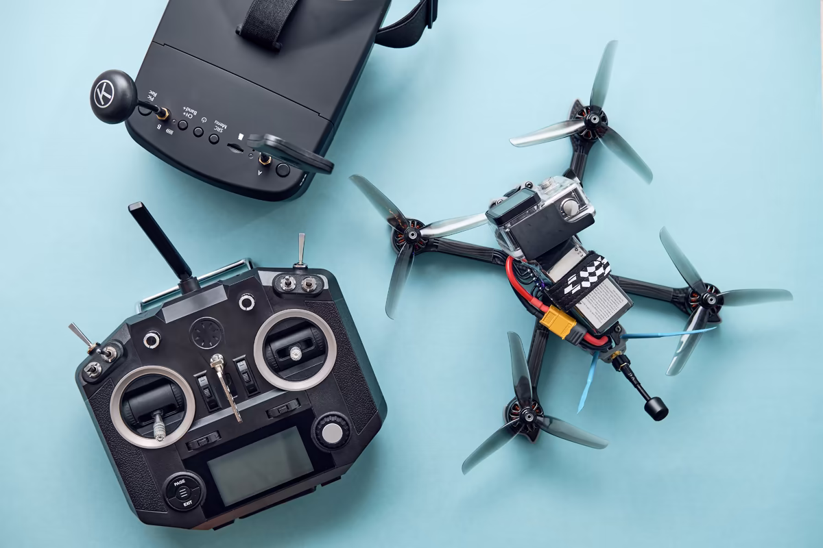Flat lay composition of drone racing gear on a light blue surface, including a quadcopter with an attached action camera, a multi-channel radio transmitter, and a black video headset.
