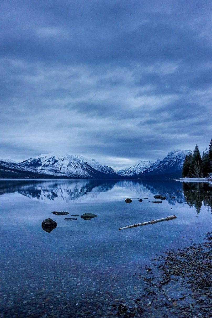 A vertical landscape shot of a still alpine lake with a pebbled shoreline, smooth rocks, and a piece of driftwood in the shallow foreground, perfectly mirroring snow-capped mountains under an overcast blue sky.