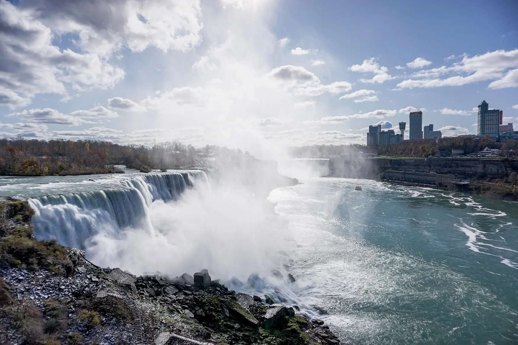 A side view of the wide American Falls at Niagara rushing over the crest, creating a heavy plume of white spray over the turquoise river below, with modern hotel towers visible on the distant horizon.