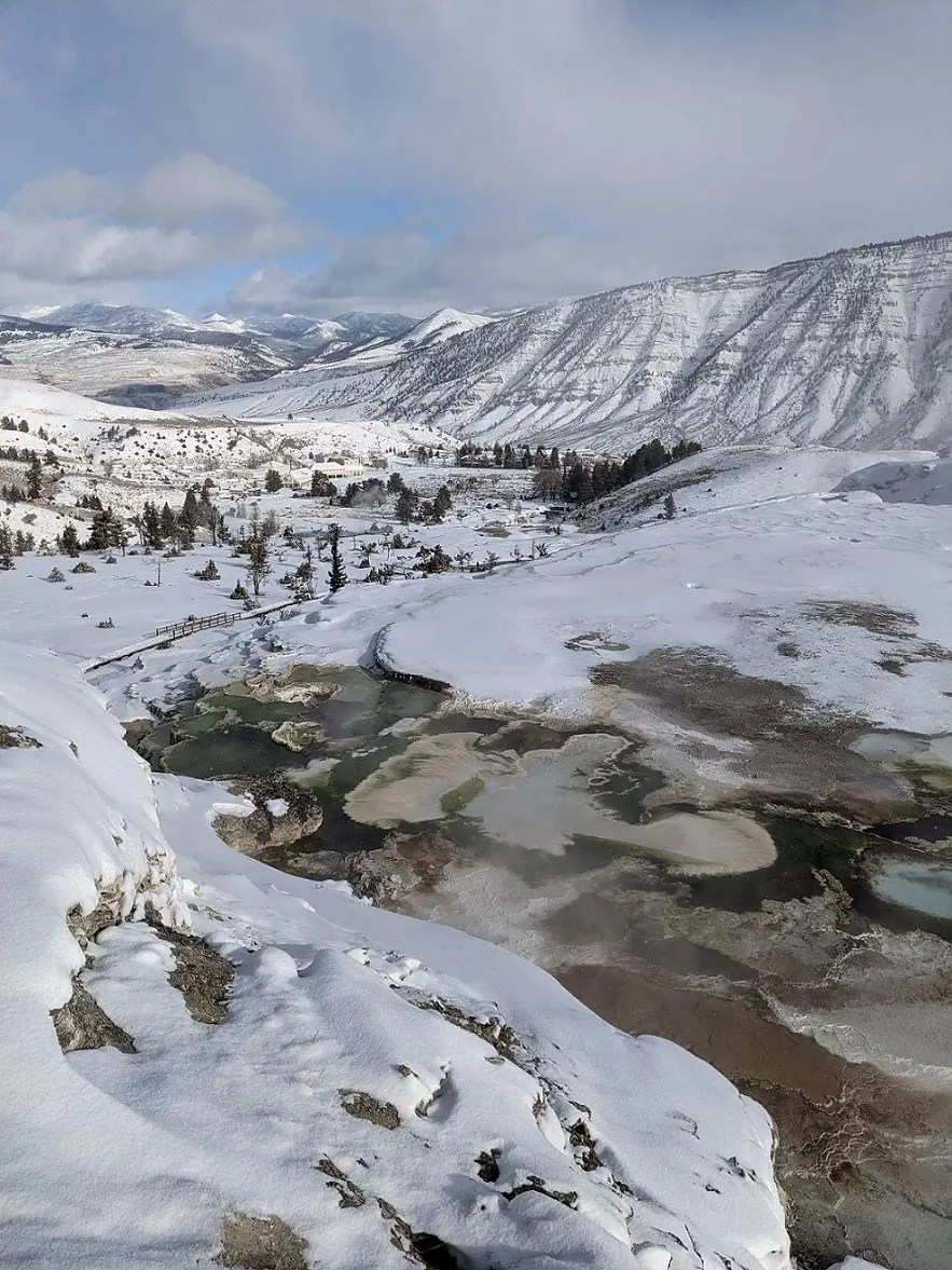 A high-angle view of snow-blanketed travertine terraces at Mammoth Hot Springs, showing pockets of exposed mineral pools steaming against a winter backdrop of snow-dusted valleys and distant ridges.